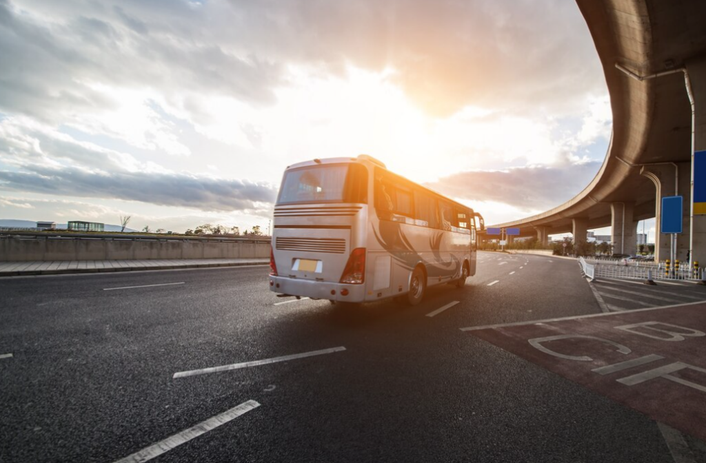 A white bus travels on an open road near a curved overpass under the cloudy sky and sunset