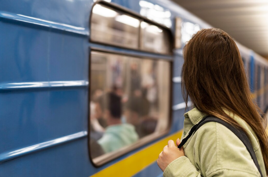 A woman with long brown hair stands close to a blue train, looking through the window at the passengers inside